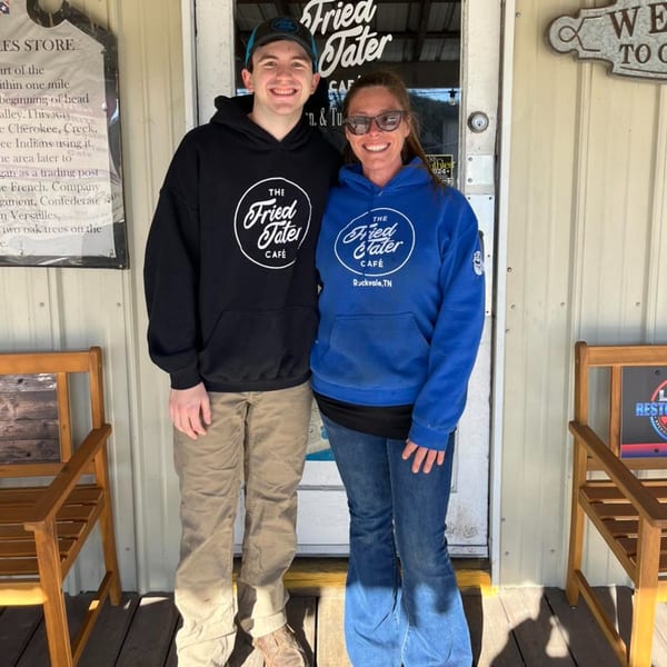 Two smiling people, a man and a woman, wearing Fried Tater Cafe hoodies stand outside the restaurant.