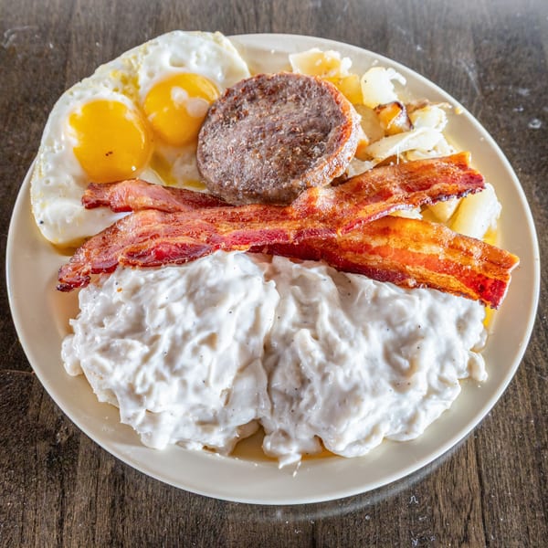 A southern breakfast plate with fried eggs, sausage, bacon, home fries, and biscuits with gravy.