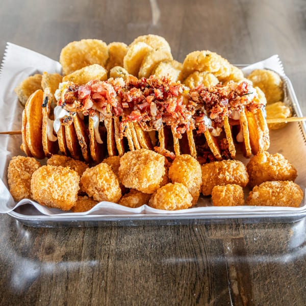 A spiral-cut potato skewer topped with bacon and sour cream, surrounded by crispy tater tots.